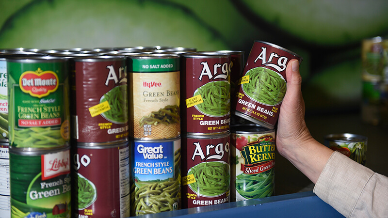 Person's hand reaches to put a can of green beans on a shelf with other green beans on a shelf. The cans all have labels from different brands, suggesting they were donated to the food shelf rather than purchased like in a store.